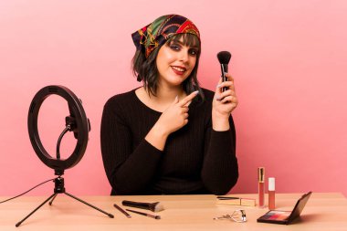 Makyaj yapan genç bir kadın pembe arka planda izole edilmiş makyaj dersi veriyor. Gülümsüyor ve işaret ediyor. Boş alanda bir şey gösteriyor..