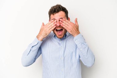 Beyaz arka planda soyutlanmış beyaz tenli genç bir adam elleriyle gözlerini kapatıyor, geniş bir gülümsemeyle bir sürpriz bekliyor..