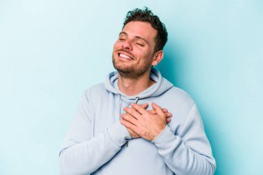 Kafkasyalı genç adam mavi arka planda izole olmuş gülüyor, elini yüreğine koyuyor, mutluluk kavramını koruyor..