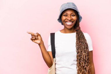 Genç Afrikalı Amerikalı bir kadın pembe arka planda izole bir şekilde kumsala gidiyor gülümsüyor ve boş alanda bir şeyler gösteriyor..