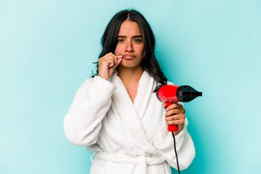 Kurutma makinesini tutan genç İspanyol kadın mavi arka planda izole edilmiş ve parmakları dudaklarında bir sır saklıyor..
