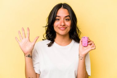 Pembe arka planda izole edilmiş anahtar taşıyan genç İspanyol kadın gülümseyerek beş numarayı parmaklarıyla gösteriyor..