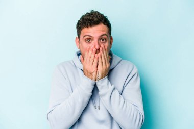 Mavi arka planda izole edilmiş genç beyaz adam şok olmuş, ağzı ellerle kaplanmış, yeni bir şey keşfetmeye hevesli..