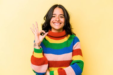 Sarı arka planda izole edilmiş genç İspanyol kadın bir göz kırptı ve elleriyle iyi bir jest yaptı..