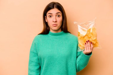 Latin kökenli genç bir kadın, bej arka planda izole edilmiş bir çanta tutuyor omuzlarını silkiyor ve gözleri açık bakıyor..