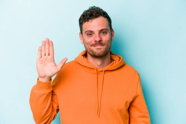 Mavi arka planda izole edilmiş genç beyaz adam gülümseyerek beş numarayı parmaklarıyla gösteriyor..