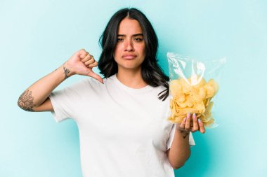Mavi arka planda izole edilmiş bir çanta dolusu cipsi tutan genç İspanyol kadın hoşlanmadığı bir hareket gösteriyor. Anlaşmazlık konsepti.