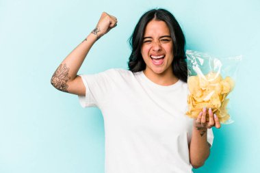 Mavi arka planda bir çanta dolusu cips tutan genç İspanyol kadın zaferden sonra yumruğunu kaldırıyor, kazanan konsept.