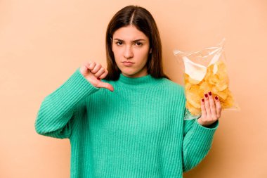 Latin kökenli genç bir kadın bej arka planda izole edilmiş bir paket cipsi tutuyor. Sevilmeyen bir jest gösteriyor. Anlaşmazlık konsepti.