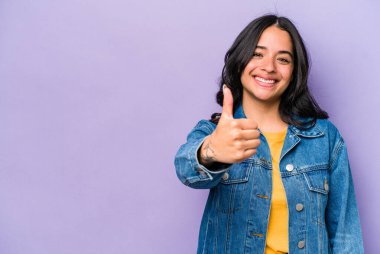 Mor arka planda izole edilmiş genç İspanyol kadın gülümsüyor ve başparmağını kaldırıyor.