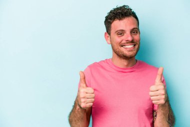 Mavi arka planda izole edilmiş genç beyaz adam gülümsüyor ve başparmağını kaldırıyor.