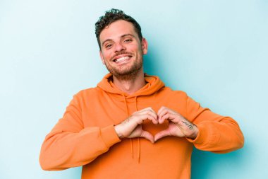 Mavi arka planda izole edilmiş genç beyaz adam gülümsüyor ve elleriyle kalp şeklini gösteriyor..
