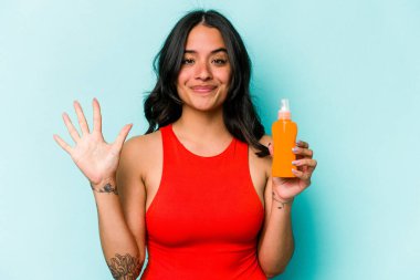Mavi arka planda güneş kremi tutan genç İspanyol kadın gülümseyerek beş numarayı parmaklarıyla gösteriyor..
