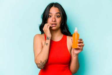 Mavi arka planda güneş kremi tutan genç İspanyol kadın tırnaklarını yiyor, gergin ve çok endişeli..