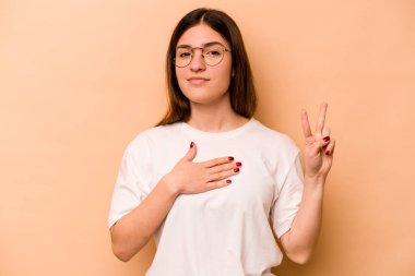 Genç İspanyol kadın bej arka planda izole edilmiş yemin ediyor, elini göğsüne koyuyor..