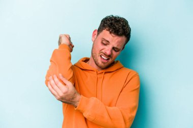 Kafkasyalı genç adam mavi arka planda izole edilmiş dirsek masajı yapıyor, kötü bir hareketin ardından acı çekiyor..