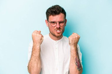 Mavi arka planda izole edilmiş genç beyaz adam yumruğunu kameraya gösteriyor, agresif yüz ifadesi var..