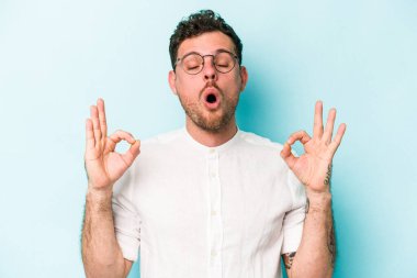 Mavi arka planda izole edilmiş genç beyaz adam yorucu bir günün ardından rahatlıyor. Yoga yapıyor..