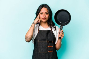 Young hispanic cooker woman holding frying pan isolated on blue background pointing temple with finger, thinking, focused on a task.