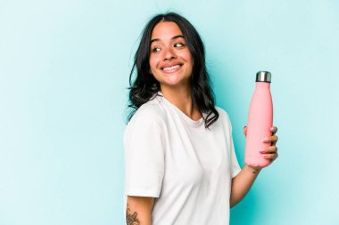 Mavi arka planda termosu olan genç İspanyol kadın gülümsüyor, neşeli ve keyifli görünüyor..