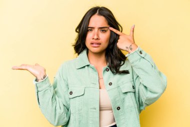 Sarı arka planda izole edilmiş genç İspanyol kadın elinde bir ürün tutuyor..