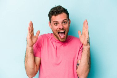 Mavi arka planda izole edilmiş genç beyaz adam hoş bir sürpriz alıyor, heyecanlı ve el kaldırıyor..