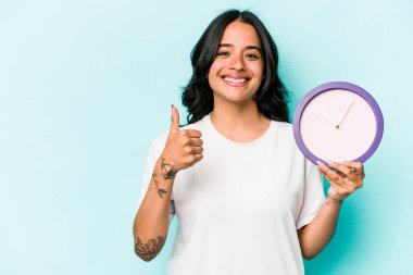 Mavi arka planda bir saat tutan genç İspanyol kadın gülümsüyor ve başparmağını kaldırıyor.