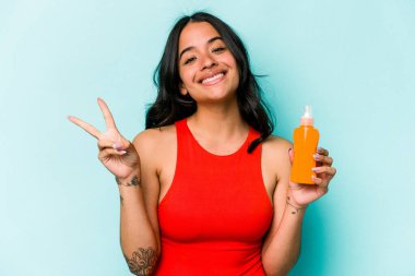 Mavi arka planda güneş kremi tutan genç İspanyol kadın neşeli ve kaygısız parmaklarıyla barış sembolü gösteriyor..