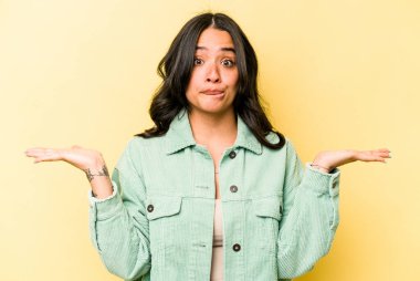 Sarı arka planda izole edilmiş genç İspanyol kadın. Kafası karışmış ve omuz silkiyor. Kopya alanını tutmak için..