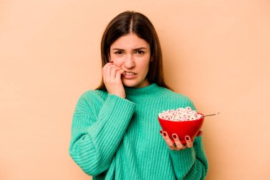 Latin kökenli genç bir kadın bej arka planda izole edilmiş mısır gevreği yiyor tırnaklarını yiyor, gergin ve çok endişeli..