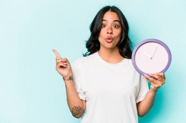 Mavi arka planda izole edilmiş bir saat tutan genç İspanyol kadın yan tarafı gösteriyor.