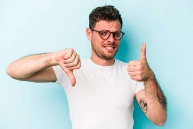 Kafkasyalı genç adam mavi arka planda izole edilmiş, baş parmağını kaldırıyor, zor seçim konsepti.