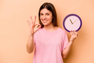 Latin kökenli genç bir kadın bej arka planda izole edilmiş bir saat tutuyor. Neşeli ve kendinden emin..
