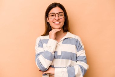 Latin kökenli genç bir kadın bej arka planda izole edilmiş mutlu ve kendinden emin gülümsüyor, elleriyle çenesine dokunuyor..