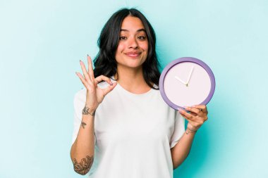 Mavi arka planda bir saat tutan genç İspanyol kadın neşeli ve kendinden emin..