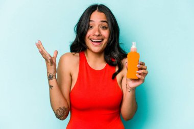 Mavi arka planda güneş kremi tutan genç İspanyol kadın hoş bir sürpriz yaşıyor, heyecanlı ve el kaldırıyor..