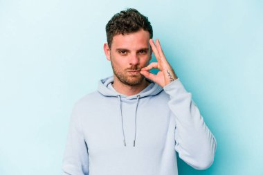 Genç beyaz adam mavi arka planda izole edilmiş ve parmakları dudaklarında bir sır saklıyor..
