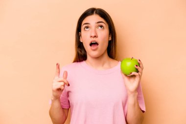 Elinde bej arka planda izole edilmiş bir elma tutan genç İspanyol kadın ağzı açık bir şekilde üst tarafı işaret ediyor..