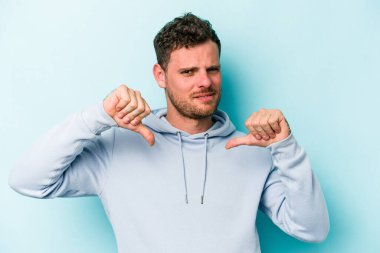 Mavi arka planda izole genç beyaz adam baş parmağını aşağı gösteriyor, hayal kırıklığı konsepti.