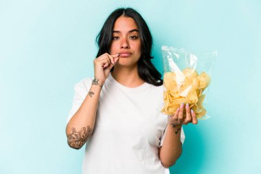 Mavi arka planda izole edilmiş bir paket cipsi tutan genç İspanyol kadın dudaklarında parmaklarıyla bir sır saklıyor..