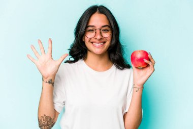 Mavi arka planda izole edilmiş bir elma tutan genç İspanyol kadın gülümseyerek beş numarayı parmaklarıyla gösteriyor..