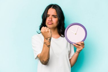 Mavi arka planda bir saat tutan genç İspanyol kadın kameraya yumruk gösteriyor, agresif yüz ifadesi..