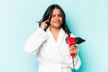 Elinde kurutucu tutan genç İspanyol kadın mavi arka planda izole edilmiş kulakları elleriyle kapatıyor..