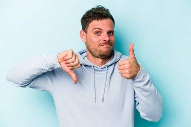 Kafkasyalı genç adam mavi arka planda izole edilmiş, baş parmağını kaldırıyor, zor seçim konsepti.