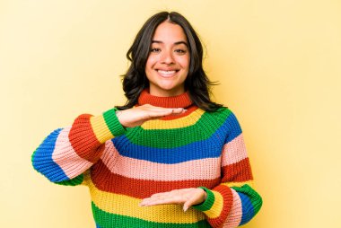 Sarı arka planda izole edilmiş genç İspanyol kadın iki eliyle bir şey tutuyor, ürün sunumu..