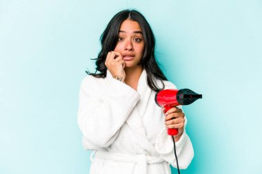 Mavi arka planda izole edilmiş, tırnak yiyen, gergin ve endişeli genç İspanyol kadın kurutucuyu tutuyor..