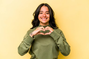 Sarı arka planda izole edilmiş genç İspanyol kadın gülümsüyor ve elleriyle kalp şeklini gösteriyor..