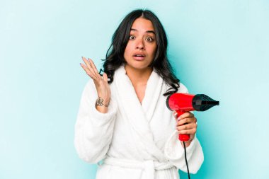 Mavi arka planda kurutma makinesini tutan genç İspanyol kadın şaşırdı ve şok oldu..