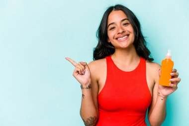 Mavi arka planda güneş kremi tutan genç İspanyol kadın gülümsüyor ve boş alanda bir şey gösteriyor..