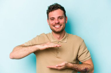 Mavi arka planda izole genç beyaz adam iki eliyle bir şey tutuyor, ürün sunumu..